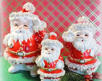 May include: Three vintage ceramic Santa Claus figurines with white fur trim and red and gold accents. The figurines are arranged in a size order from largest to smallest.