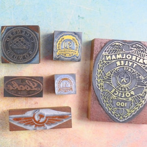 May include: A collection of six vintage rubber stamps. The stamps are made of wood and metal and feature various designs, including a police badge, a company logo, and a 50th anniversary logo.