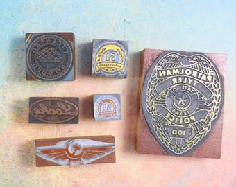 May include: A collection of six vintage rubber stamps. The stamps are made of wood and metal and feature various designs, including a police badge, a company logo, and a 50th anniversary logo.