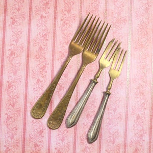May include: Four antique gold forks on a pink and gold patterned background. The forks are arranged in a diagonal line, with the largest fork at the top and the smallest fork at the bottom.
