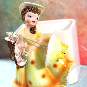 May include: A vintage ceramic figurine of a woman in a yellow dress and white hat holding a basket of flowers and an umbrella. The figurine is a planter with a white base.