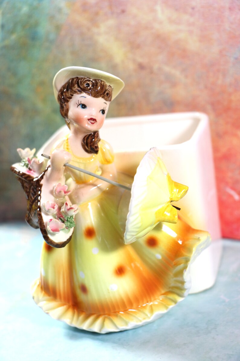 May include: A vintage ceramic figurine of a woman in a yellow dress and white hat holding a basket of flowers and an umbrella. The figurine is a planter with a white base.