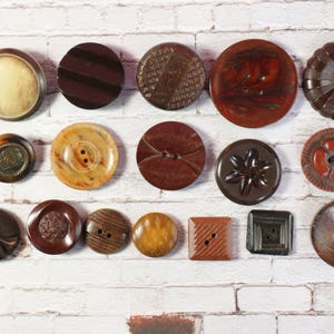 May include: Assortment of vintage buttons in various colors, patterns, and materials, including brown, black, white, and red. The buttons are arranged in rows on a white brick background.