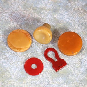 May include: A collection of five vintage buttons and fasteners including two round amber-colored buttons with smooth surfaces, one light tan domed button, one bright red circular button with a center hole, and one red keyhole-shaped fastener.