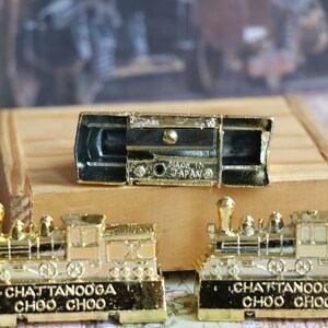 Op de afbeelding: Twee goudkleurige speelgoedtrein potloodpunten met de woorden "Chattanooga Choo Choo" op de basis. De puntenslijpers zijn open, waardoor het zwarte slijpmechanisme zichtbaar is. De bovenkant van de puntenslijpers heeft een goudkleurige speelgoedtrein.