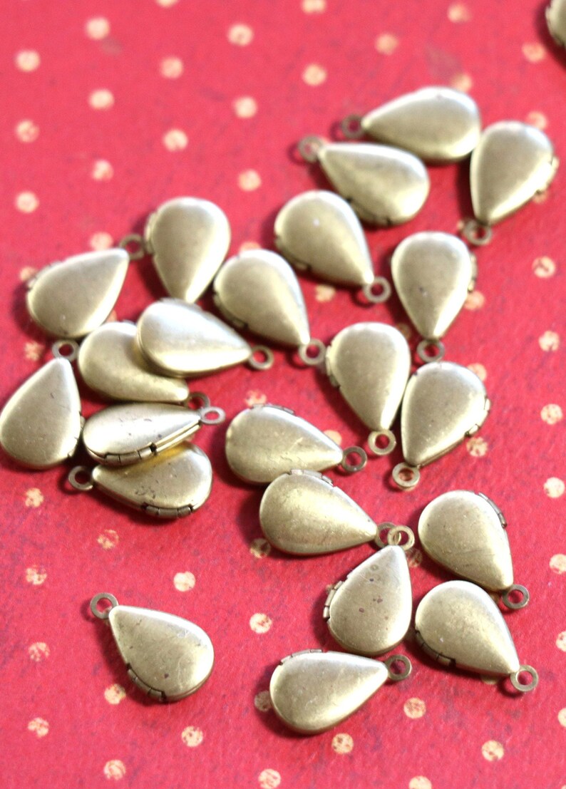 May include: A group of 20 gold-toned teardrop-shaped metal charms with a hinged opening. The charms are scattered on a red background with white polka dots.