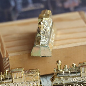 Op de afbeelding: Drie gouden miniatuurtreinlocomotieven. De voorste locomotief is naar voren gericht en de andere twee zijn naar de kijker gericht. De locomotieven staan op een houten oppervlak.