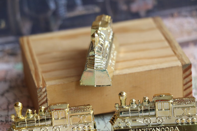 Op de afbeelding: Drie gouden miniatuurtreinlocomotieven. De voorste locomotief is naar voren gericht en de andere twee zijn naar de kijker gericht. De locomotieven staan op een houten oppervlak.