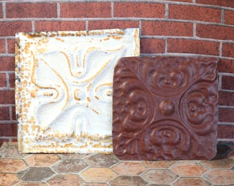 May include: Two decorative square tiles. One is off-white with a floral design and aged, distressed edges. The other is a deep brown with a raised, ornate pattern. Both tiles are leaning against a red brick wall.