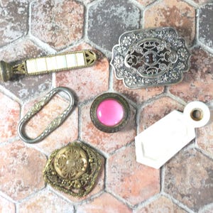 May include: Five antique metal and ceramic decorative pieces on a hexagonal tile background. The pieces include a long, ornate, silver-colored handle, a silver-colored oval ring, a small, round, pink-colored button with a silver-colored rim, a small, white, rectangular tag with a hole in the top, and a small, gold-colored, ornate button.