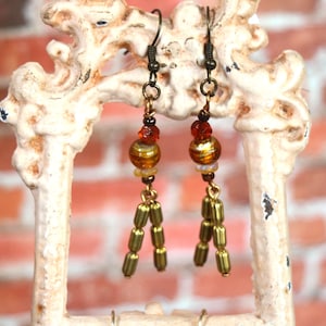 May include: A pair of dangle earrings with a vintage aesthetic. Each earring features a dark metal hook, a cluster of orange and gold beads, and a cascade of gold-colored cylindrical beads. The earrings are displayed against a white, ornate frame.