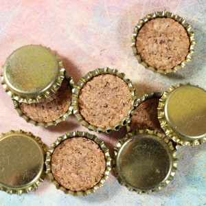 May include: A group of eight gold bottle caps with cork inserts. The bottle caps are arranged in a random pattern on a textured, multi-colored surface.