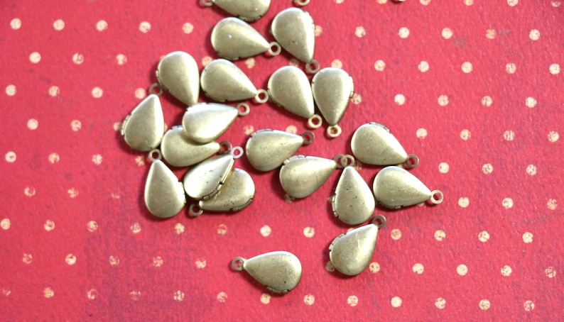 May include: A group of gold teardrop-shaped charms with a loop at the top. The charms are scattered on a red background with white polka dots.
