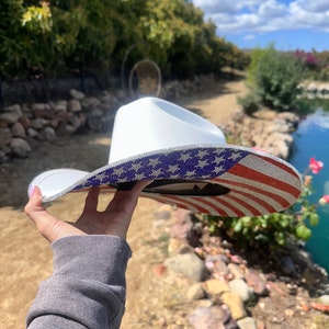K&ouml;nnte beinhalten: Ein wei&szlig;er Cowboyhut mit einem glitzernden amerikanischen Flaggenmuster. Der Hut wird von einer Hand hochgehalten.