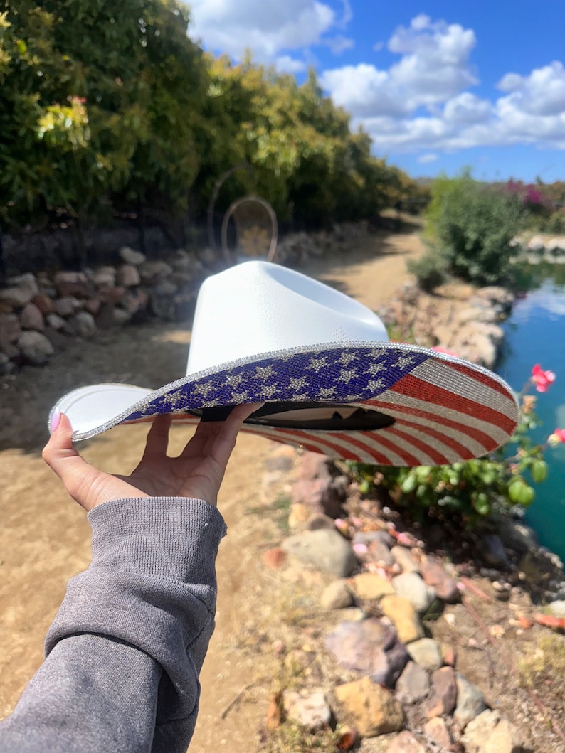 K&ouml;nnte beinhalten: Ein wei&szlig;er Cowboyhut mit einem glitzernden amerikanischen Flaggenmuster. Der Hut wird von einer Hand hochgehalten.