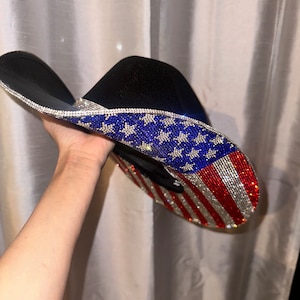 K&ouml;nnte beinhalten: Ein schwarzer Cowboyhut mit einem Strass-amerikanischen Flaggenmuster. Der Hut ist mit blauen Sternen und roten und wei&szlig;en Streifen verziert.