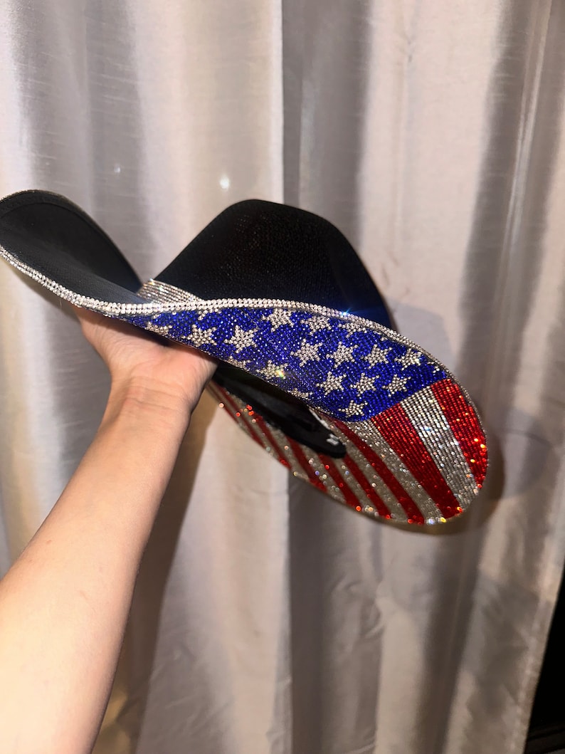 K&ouml;nnte beinhalten: Ein schwarzer Cowboyhut mit einem Strass-amerikanischen Flaggenmuster. Der Hut ist mit blauen Sternen und roten und wei&szlig;en Streifen verziert.
