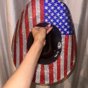 K&ouml;nnte beinhalten: Ein Cowboyhut mit einem rot-wei&szlig;-blauen amerikanischen Flaggenmuster, das mit Strasssteinen besetzt ist. Der Hut ist innen schwarz und hat ein schwarzes Band mit dem Text "XXL" darauf.