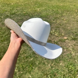 May include: A white cowboy hat with a silver rhinestone trim. The hat is held in a hand with the brim facing the camera.