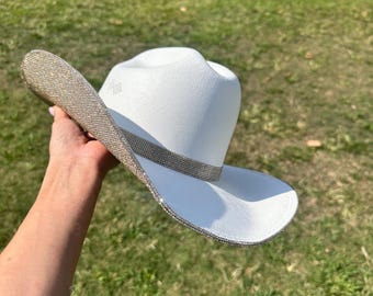 Puede incluir: Un sombrero vaquero blanco con un borde de pedrería plateada. El sombrero se sostiene en una mano con la ala mirando hacia la cámara.