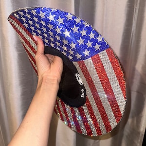 K&ouml;nnte beinhalten: Ein rot-wei&szlig;-blauer Cowboyhut, der mit Strasssteinen besetzt ist und das amerikanische Flaggenmuster zeigt. Der Hut wird von einer Hand gehalten.