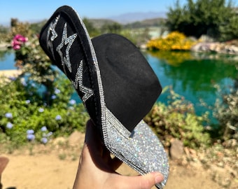 Puede incluir: Un sombrero vaquero negro con un diseño de estrellas plateadas alrededor del borde. El sombrero está decorado con diamantes de imitación.
