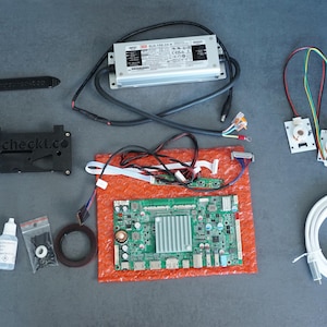 Könnte beinhalten: Eine Ansammlung elektronischer Komponenten, darunter eine Leiterplatte, ein Netzteil und verschiedene Anschlüsse, angeordnet auf einer grauen Oberfläche. Die Komponenten sind in verschiedenen Farben gehalten, darunter Grün, Schwarz und Weiß. Der Text "eyecheckt.co" ist auf einem schwarzen Bauteil sichtbar.