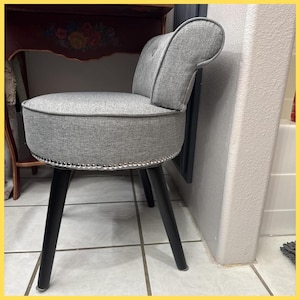 May include: A gray upholstered vanity chair with a rounded seat and backrest. The chair has a silver studded trim and black tapered legs. The chair is placed on a tiled floor.