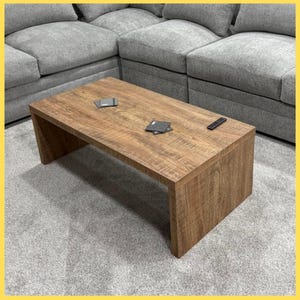 May include: A rectangular wooden coffee table with a natural wood grain finish. The table has a simple, modern design with a solid base. Small items, including coasters and a remote control, are on the table's surface. The table is in front of a gray sectional sofa.