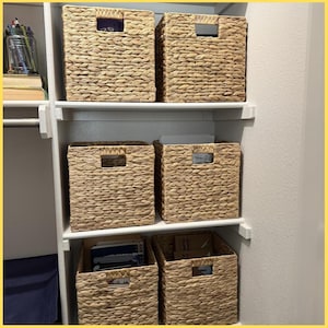 May include: Six woven storage baskets with rectangular cut-out handles are arranged on white shelves inside a closet. The baskets are made of a light brown, natural material and are filled with books and other items.