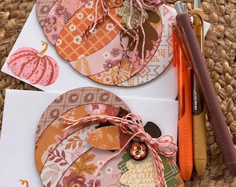 May include: Two handmade greeting cards featuring a large pumpkin shape made of layered patterned paper in shades of orange, pink, and brown. Each card is decorated with twine bows, paper leaves, and a small button. Three pens are placed beside them.