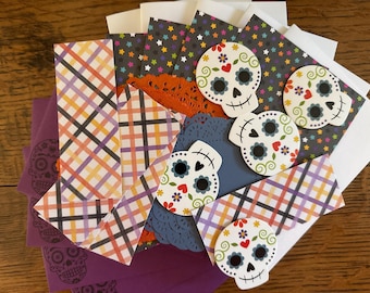 May include: A collection of stationery items featuring white sugar skull cutouts with colorful floral details, purple envelopes with skull outlines, plaid patterned paper strips, and dark paper sheets with a multicolored star print pattern.
