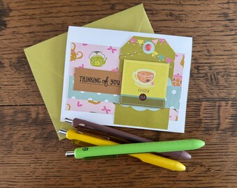 May include: A handmade greeting card with a tag that says "Ginger Tea" and "Thinking of You". The card is decorated with pink and blue patterned paper and a yellow envelope. Several pens in brown, yellow, and green are in the foreground.