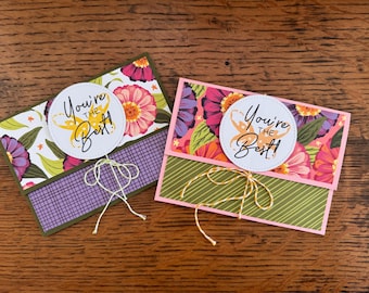 May include: Two floral greeting cards with the text "You're the Best!" on white circular labels. One card has a purple and green color scheme, the other has pink and green. Both cards are tied with twine.