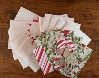 May include: A collection of Christmas-themed stationery. Includes white envelopes with red reindeer, patterned cards with candy cane stripes, floral designs, and gold glitter reindeer cutouts. The items are arranged on a wooden surface.