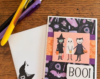 May include: A Halloween-themed greeting card with a witch, ghost, and vampire illustration, with the word "BOO!". The card is paired with a blank envelope featuring a witch hat graphic and several pens.