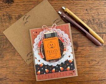 May include: A Halloween-themed greeting card with a tag that says "Treats" and a jack-o'-lantern. The card is decorated with a white doily, candy corn, and a brown envelope. Two pens are on the wooden surface.