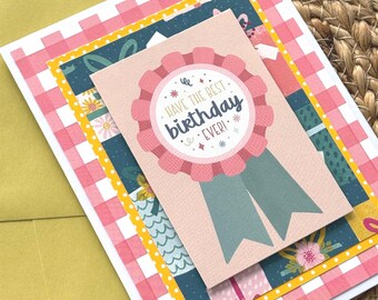 May include: A rectangular greeting card featuring a layered design with a pink and white checkered pattern, a dark teal floral panel, and a central light pink rosette badge. The badge displays the text HAVE THE BEST birthday EVER! in dark blue.