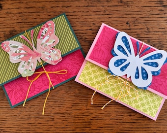 May include: Two handmade greeting cards with butterfly embellishments. One card features a pink floral pattern with a green striped flap and a pink and floral butterfly. The other card has a pink floral pattern with a yellow geometric flap and a white butterfly with blue polka dots.