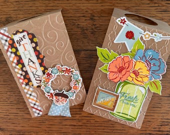 May include: Two brown paper gift bags with embossed patterns. The left bag features a plaid strip, a tag reading "give THANKS," a floral wreath, and an acorn. The right bag displays a floral bouquet in a green jar, a "Fresh FOR YOU" label, and a "PEARS" sign.