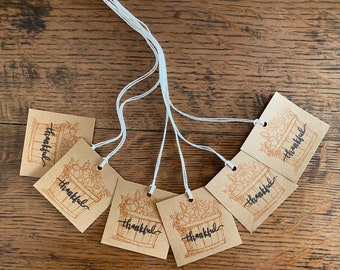 May include: Six brown paper gift tags with white twine. Each tag features a hand-drawn illustration of a basket overflowing with pumpkins and the word "thankful" written in black calligraphy.