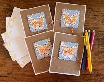 May include: Four brown cardstock cards with white borders and scalloped edges. Each card features a blue and white patterned square with a yellow and orange butterfly cutout. The butterflies have red and white accents. The cards are arranged in a row on a wooden surface.