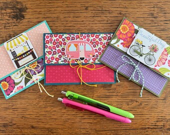 May include: Three handmade greeting cards with decorative paper and twine. One card features a lemonade stand, another a pink camper, and the third a bicycle with flowers. Two pens, one green and one pink, are in the foreground.