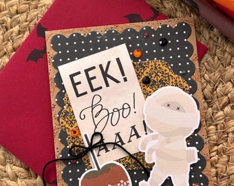 May include: A handmade Halloween greeting card featuring a scalloped black border with white polka dots, a central cream panel with the text EEK! Boo! AAA!, a cartoon mummy, and a caramel apple graphic, accented with small orange and black gems.