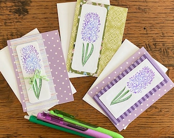 May include: Four handmade greeting cards with a purple hyacinth flower design on a white background. The cards are decorated with purple and white polka dots, stripes, and a green floral pattern. The cards are on a wooden surface with three pens.