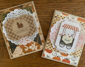 May include: Two handmade Halloween greeting cards. The first card features a brown and white paper with a white doily and a brown label with a house and the text "happy halloween". The second card features a brown and orange paper with a white doily and a brown label with a black and white illustration of a sweets cart with the text "Sweets".