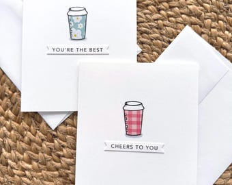 May include: Two white greeting cards with matching envelopes. One card features a light blue floral coffee cup illustration with the text YOU'RE THE BEST. The other card displays a red gingham coffee cup illustration with the text CHEERS TO YOU.