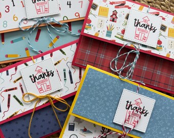 May include: Four handmade school-themed gift card holders. Each features a patterned design with school supplies, numbers, or plaid. A white square with the word "thanks" and a schoolhouse illustration is tied with twine.