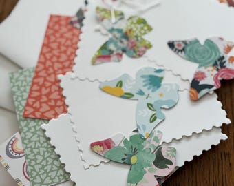 May include: A collection of handmade greeting cards with butterfly cutouts. The butterflies are made from patterned paper in various colors and designs, including floral and rainbow patterns. The cards have a scalloped edge and are stacked on a wooden surface.