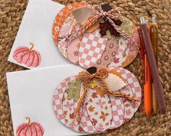 May include: Two white envelopes featuring a pink pumpkin illustration, each topped with a layered paper pumpkin cutout. The cutouts display checkered and leaf patterns in orange, pink, and brown, accented with twine bows and felt leaves, alongside three pens.